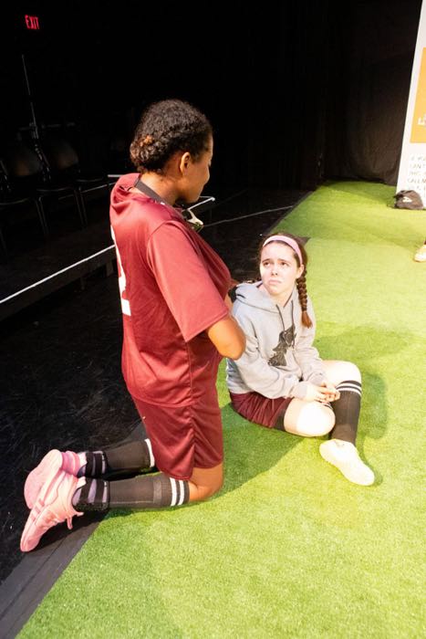 Player number two kneels next to another female soccer player who is sitting on the ground looking very sad.